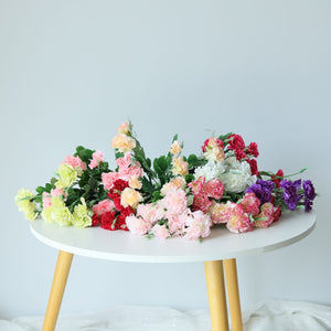 A bunch of artificial carnation spray silk flowers displayed on a white table with yellow legs against a neutral background.
